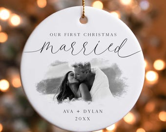 May include: White ceramic Christmas ornament with a black and white photo of a couple kissing. The text "Our First Christmas Married" is written in black script above the photo. The couple's names "Ava + Dylan" and the year "20XX" are written below the photo.