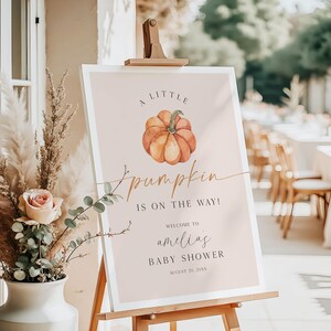 May include: A baby shower welcome sign on an easel. The sign has a light pink background with a watercolor pumpkin illustration. Text reads "A Little Pumpkin is on the Way!" and welcomes guests to Amelia's baby shower.