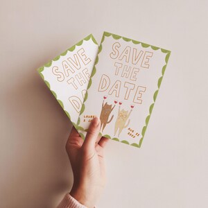 May include: Two white "Save the Date" cards with gold lettering and a green scalloped border. The cards feature illustrations of cats toasting with wine glasses. The date "Aug 11 20XX" is printed on the card.