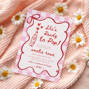 May include: A baby shower invitation with a pink gingham background and red accents. The invitation reads "She's Ready to Pop!" and includes details for the event, such as the date, time, and location. White daisies are scattered around the invitation.