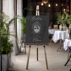 May include: A dark gray sign on a wooden easel with the text "Welcome" and "As we mourn Olivia's 20's". The sign features a portrait in an ornate frame, set against a skull design.