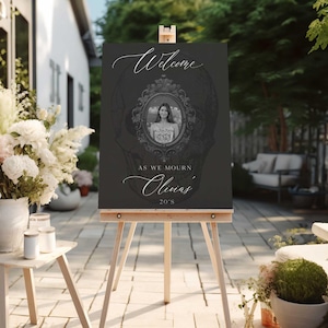 May include: A dark gray welcome sign on a wooden easel. The sign features the word "Welcome" in elegant script, a framed portrait, and the text "As We Mourn Olivia's 20's". The background has a skull design.