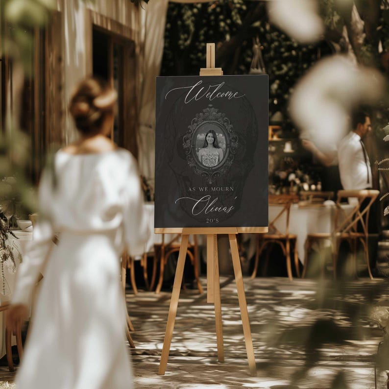 May include: A dark gray welcome sign on a wooden easel. The sign features the word "Welcome" in elegant script, a framed portrait, and the text "As we mourn Olivia's 20's."