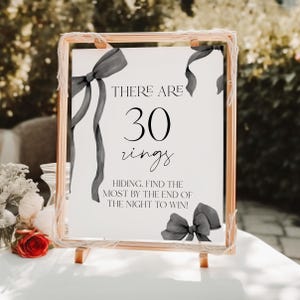 May include: A white sign with black text and gray ribbon bows. The sign reads "THERE ARE 30 rings HIDING. FIND THE MOST BY THE END OF THE NIGHT TO WIN!"