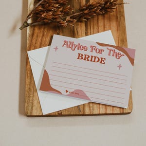 May include: A light pink advice card for the bride with the text "Advice For The Bride" in a retro font. The card has a white envelope and is decorated with brown and pink abstract shapes and star accents. The words "with love" are at the bottom.