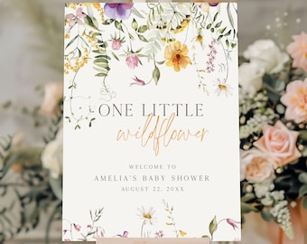 Puede incluir: Un letrero de bienvenida para baby shower con ilustraciones florales. El letrero presenta el texto "ONE LITTLE wildflower" en una fuente de escritura, con texto adicional debajo. El letrero está decorado con flores de acuarela en tonos amarillos, morados y rosas.