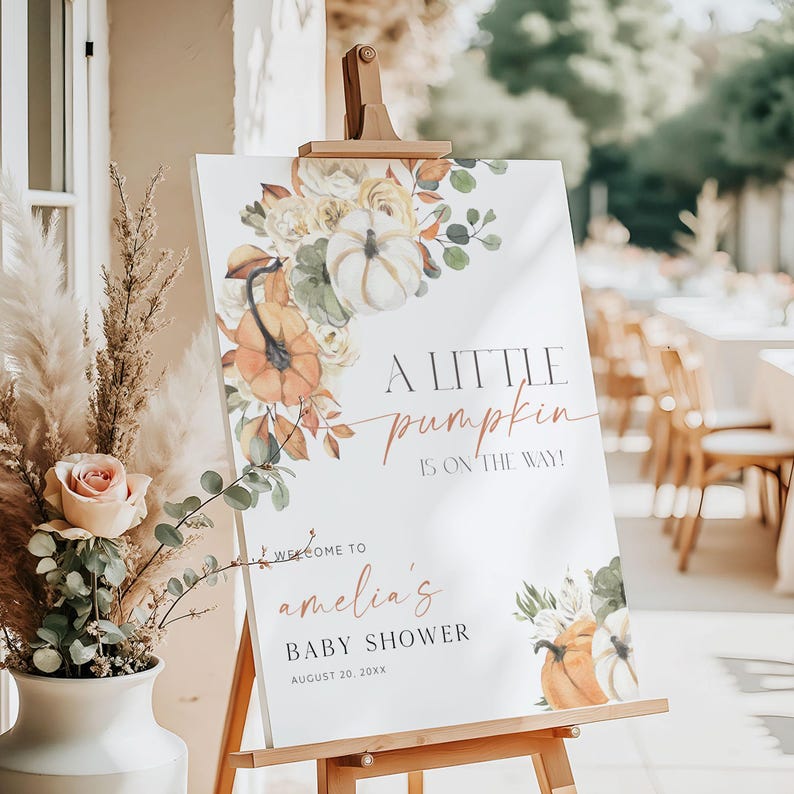 May include: A white sign on a wooden easel, decorated with pumpkins and floral arrangements. The sign reads "A LITTLE pumpkin IS ON THE WAY!" and welcomes guests to "amelia's BABY SHOWER" on August 20, 20XX.