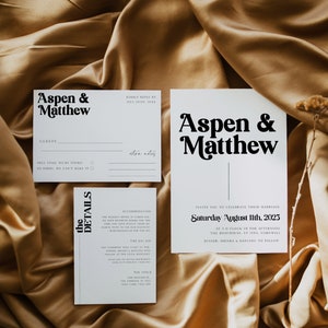 May include: White wedding invitation cards with black text. The top card says "Aspen & Matthew" in a bold, retro font. The bottom card has the details of the wedding, including the date, time, and location. The text reads "Invite you to celebrate their marriage Saturday August 11th, 2023 at 3 o'clock in the afternoon The Boathouse, St Ives, Cornwall Dinner, drinks & dancing to follow."