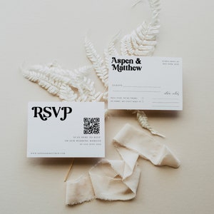 May include: A white RSVP card with a black QR code and the text "SCAN HERE TO RSVP ON OUR WEDDING WEBSITE BY JULY 20TH, 20XX". The card also includes the text "RSVP" in a stylized font and the website address "WWW.ASPENANDMATTHEW.COM".