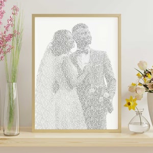 May include: A framed art print featuring a silhouette of two people embracing, filled with song lyrics in a black and white design. The artwork is encased in a gold-colored frame and rests on a wooden surface. Decorative flowers in vases are on either side.