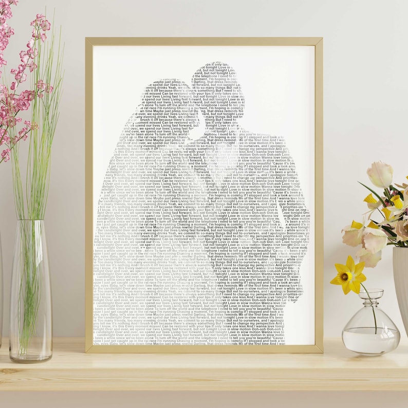 May include: A framed art print featuring a silhouette of two people embracing, filled with song lyrics in a black and white design. The artwork is encased in a gold-colored frame and rests on a wooden surface. Decorative flowers in vases are on either side.