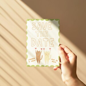 May include: A rectangular "Save the Date" card with a green scalloped border. The card features two cartoon cats holding wine glasses. The text "SAVE THE DATE" is at the top, with names and a date below.