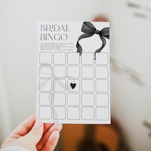 May include: A black and white bridal bingo card with a black bow at the top. The card has 25 squares with a black heart in the center square. The text on the card reads "Bridal Bingo" and "Fill the squares with gifts you think the bride-to-be will receive. As the gifts are opened, mark off any matches on your card. When you get 5 in a row, tell bingo!"