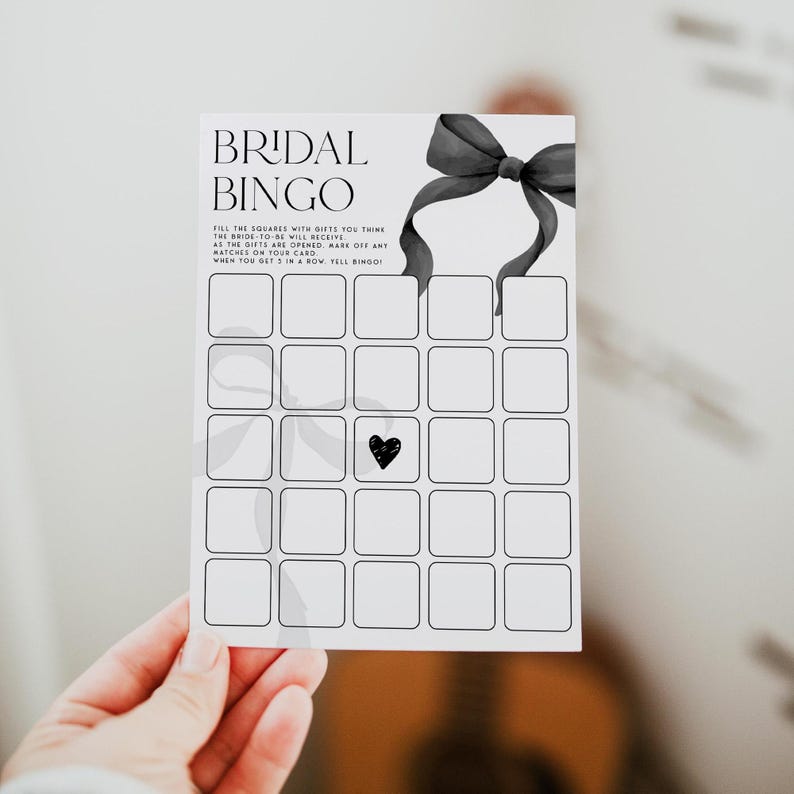 May include: A black and white bridal bingo card with a black bow at the top. The card has 25 squares with a black heart in the center square. The text on the card reads "Bridal Bingo" and "Fill the squares with gifts you think the bride-to-be will receive. As the gifts are opened, mark off any matches on your card. When you get 5 in a row, tell bingo!"