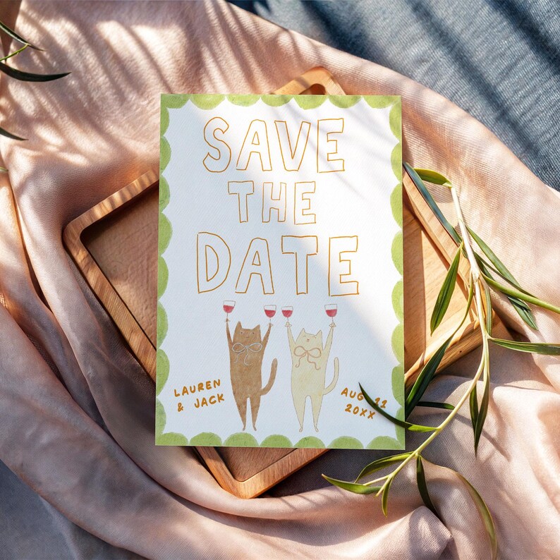 May include: A Save the Date card with gold lettering and a green scalloped border. Two cartoon cats are raising wine glasses. The card is on a wooden tray, with the names "Lauren & Jack" and the date "Aug 15 20XX" printed on it.