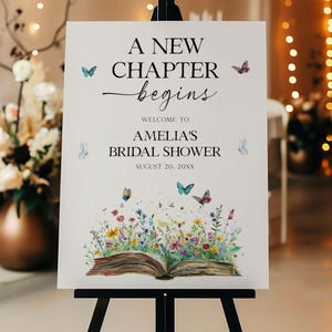 May include: A white sign on a black easel with the text "A NEW CHAPTER begins" and "WELCOME TO AMELIA'S BRIDAL SHOWER AUGUST 20. 20XX". The sign is decorated with butterflies and a watercolor illustration of an open book with flowers.