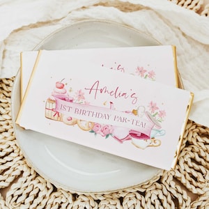 May include: A white chocolate bar wrapper with a pink and gold design. The wrapper features a watercolor illustration of teacups, flowers, and a cake with the text "Amelia's 1st Birthday Par-Tea!"
