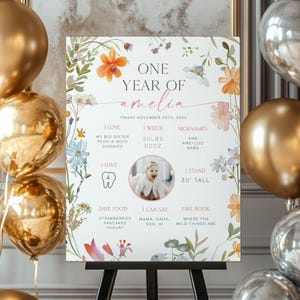 May include: A white sign with floral accents and the text "ONE YEAR OF amelia." It includes baby stats: weight, height (30 inches), favorite foods, and nicknames. The sign is on a black easel, with gold and silver balloons in the background.