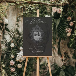 May include: A dark gray welcome sign on a wooden easel. The sign features the word "Welcome" in elegant script, a framed portrait, and the text "As we mourn Olivia's 20's."