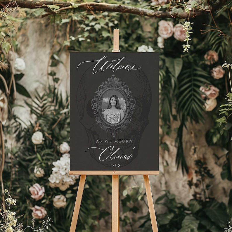 May include: A dark gray welcome sign on a wooden easel. The sign features the word "Welcome" in elegant script, a framed portrait, and the text "As we mourn Olivia's 20's."