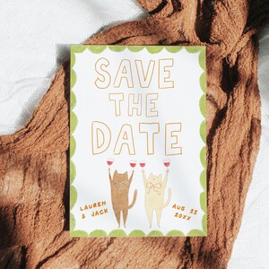 May include: A white Save the Date card with a green scalloped border. The card features two cartoon cats holding wine glasses. The text "SAVE THE DATE" is in gold, along with the names "LAUREN & JACK" and the date "AUG 22 20XX".