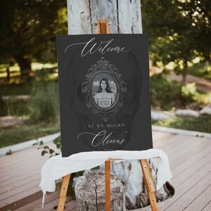 May include: A dark gray welcome sign on an easel. The sign features the word "Welcome" in elegant script, a framed photo, and the text "As we mourn Olivia's 20's". The background has a skull design.