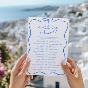 May include: A white card with blue text and a blue bow at the top. The card has the text "would she rather?" and a list of questions with two options each. The questions are about the bride's preferences, such as "Drink coffee or sip on some tea?" and "Go to the movies or watch a film at home?"