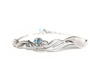 May include: A silver bracelet with a floral and wave design, adorned with several small, light blue gemstones. The bracelet is set against a white background, highlighting its intricate details and delicate craftsmanship. The bracelet appears to be made of sterling silver.