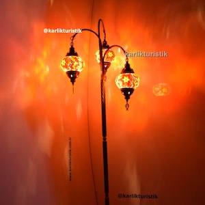 Op de afbeelding: Een drielampen vloerlamp met een gebogen, goudkleurig metalen frame en drie ronde, kleurrijke mozaïek glazen lampenkappen. De lamp is aan en geeft een warm oranje licht. De tekst "@karlikturistik" is zichtbaar op de muur achter de lamp.