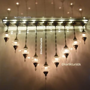 May include: A chandelier with multiple clear glass globes hanging from a metal frame. The globes are lit with warm white light bulbs. The chandelier is hanging from a ceiling with a white background. The text "karlikturistik" is visible in the image.