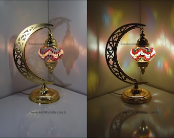 May include: A gold metal table lamp with a crescent moon shaped base and a colorful mosaic glass globe. The lamp is lit and casting a warm glow.