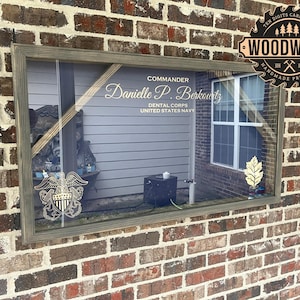 May include: A framed wooden display case with a glass front, showcasing a military emblem and text that reads "Commander Danielle P. Berkowitz, Dental Corps, United States Navy." The frame is gray, and the background is a brick wall.