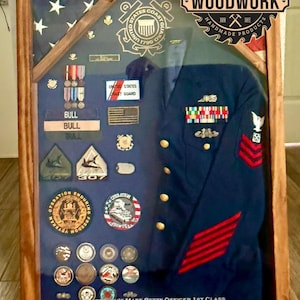 May include: A wooden shadow box displaying a U.S. Coast Guard uniform, medals, and a flag. The uniform is navy blue with gold buttons and red stripes. The box includes the text "Michael S. Bull" and dates. The frame is made of stained wood.