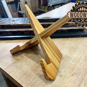 May include: A wooden phone stand with a crossed-beam design. The stand is made of light-colored wood with visible grain and a slot to hold a phone. The stand is on a wooden surface. The image also shows a wooden display case with an American flag design.