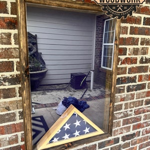 May include: A wooden frame with a glass front, displaying a folded American flag in a shadow box. The frame is mounted on a brick wall. The text "TEN DIGITS CARPENTRY WOODWORK HANDMADE PRODUCTS" is visible on a circular label.