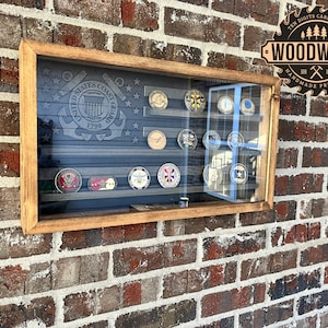 May include: A wooden shadow box with a glass front displays a collection of military challenge coins. The shadow box is mounted on a brick wall. The shadow box has a dark wood frame and a clear glass front. The coins are arranged in rows and columns. The coins are all different sizes and shapes. The coins are all made of metal and have different designs. The shadow box is labeled "WOODWORK HANDMADE PRODUCTS".