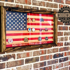 May include: A wooden shadow box with a red, white, and blue American flag background. The shadow box has a glass front and is displaying a collection of military challenge coins. The text "MSGT DAVID C THIES, US AIR FORCE" is printed on the bottom of the shadow box. The shadow box is mounted on a brick wall. The logo "WOODWORK TEN DIGITS CARPENTRY HANDMADE PRODUCTS" is visible in the upper right corner of the image.