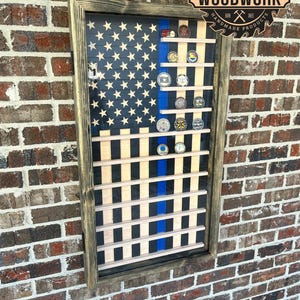 May include: A wooden shadow box with a blue and white American flag design and a thin blue line. The shadow box has slots for displaying challenge coins. The shadow box is framed in dark wood and has a glass front. The shadow box is mounted on a brick wall. The text "TEN DIGITS CARPENTRY WOODWORK HANDMADE PRODUCTS" is visible on the shadow box.
