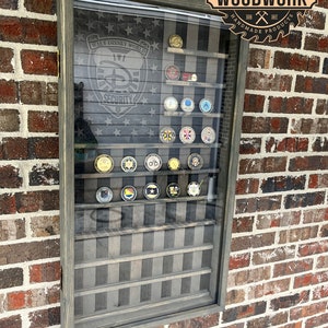 May include: A wooden shadow box display case with a black and white American flag background. The case has multiple shelves for displaying round metal coins. The case is mounted on a brick wall. The text "WALT DISNEY WORLD SECURITY" is visible on the flag. The text "TEN DIGITS CARPENTRY WOODWORK HANDMADE PRODUCTS" is visible on a circular saw blade.