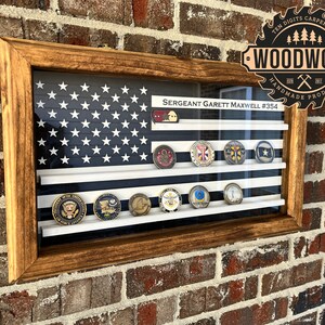 May include: A wooden shadow box with a printed American flag background and a white frame. The shadow box displays a collection of military challenge coins with the text "SERGEANT GARETT MAXWELL #354" above the coins. The shadow box is labeled "WOODWORK HANDMADE PRODUCTS" in a circular saw blade design.
