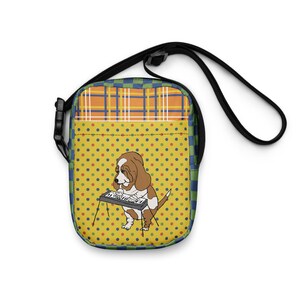 May include: A yellow and blue patterned crossbody bag with a cartoon illustration of a dog playing a keyboard.