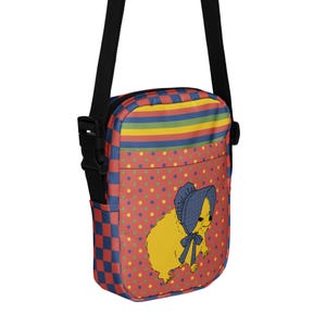 May include: A crossbody bag with a whimsical design. The bag features a red background with yellow polka dots and a cartoon illustration of a yellow creature wearing a blue bonnet. The top has rainbow stripes, and the side has a blue and red checkered pattern.