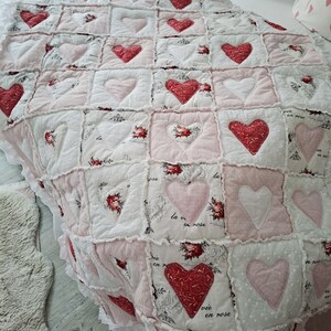 Könnte beinhalten: Eine handgefertigte Steppdecke mit Patchwork-Design. Sie zeigt rote und rosa Herzen auf weißem und rosa Hintergrund. Einige Quadrate haben florale Muster und den Ausdruck "la vie en rose". Die Steppdecke hat einen gerüschten Rand.