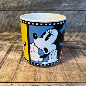 May include: A white ceramic mug with a black, white and yellow design featuring Mickey Mouse and Minnie Mouse. The design is framed with a black and white film strip.