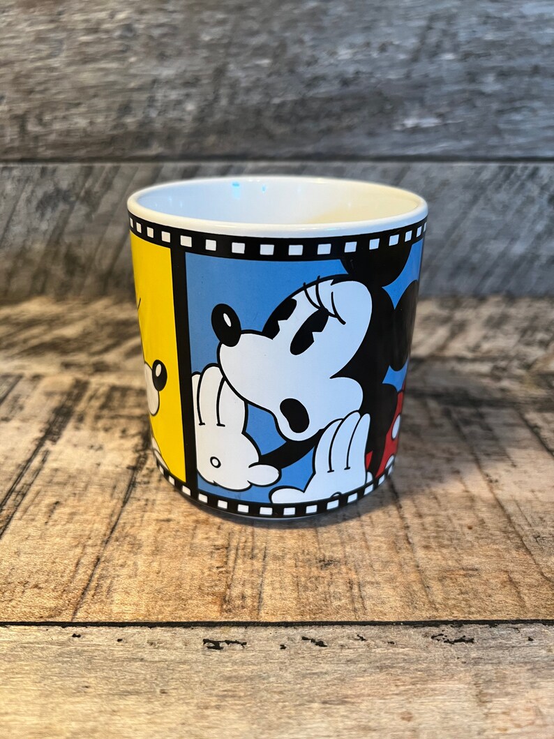 May include: A white ceramic mug with a black, white and yellow design featuring Mickey Mouse and Minnie Mouse. The design is framed with a black and white film strip.