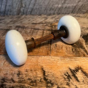May include: A vintage door knob set featuring two smooth, white, rounded ceramic knobs connected by a central, square-shaped, rusted metal spindle. The hardware shows signs of age with visible oxidation on the metal components and a glossy finish on the ceramic surfaces, creating a functional assembly for a door mechanism.