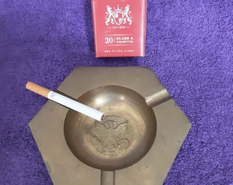 May include: A brass ashtray with a hexagonal shape and a raised eagle design in the center. A cigarette is resting on the edge of the ashtray. A pack of Pall Mall Red Filter Kings cigarettes is in the background.