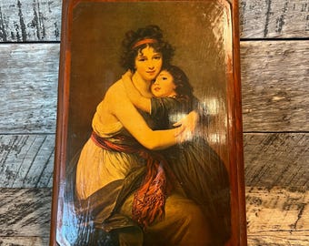 May include: A framed print of a woman and child embracing. The artwork features warm tones, with the figures in light-colored clothing and a red sash. The frame is dark wood with a metal loop for hanging, suggesting a vintage aesthetic.