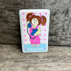 May include: A rectangular white telephone book featuring a cartoon illustration of a brown-haired child with pigtails wearing pink overalls and holding a blue telephone receiver. The background is white with small pink hearts and the text Telephone Book.