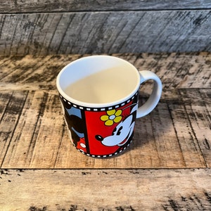 May include: A white ceramic mug with a black and red design featuring Mickey Mouse and Minnie Mouse. The mug has a black handle and a film strip border.